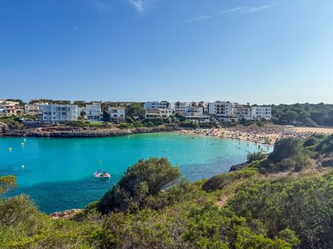 Turquoise bay of Cala Marcal beach in Porto Colom, Mallorca, with white buildings along the shore and green vegetation in foreground.