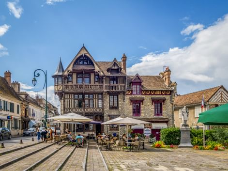 Half-timbered Maison Raccollet building in Moret-sur-Loing with outdoor café seating on a cobblestone square under blue sky.