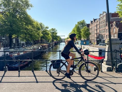 Radfahrerin auf Brücke mit Blick auf Amsterdamer Gracht mit Hausbooten, traditionellen niederländischen Gebäuden und Bäumen am Ufer.