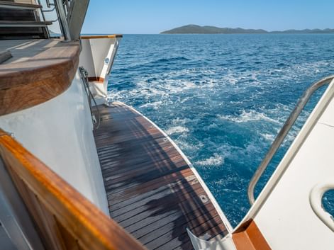 View from wooden deck of MS Macek ship showing teak planking, white railings, and blue sea with distant islands under clear sky.