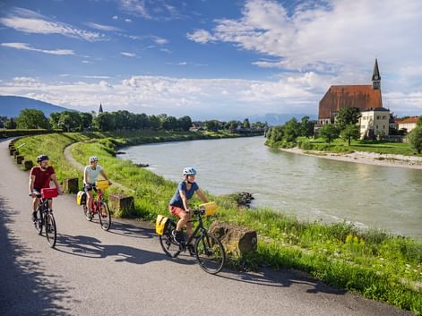 Drei Radfahrer fahren am Salzachradweg bei Laufen. Eine historische Kirche mit rotem Dach und Turm steht am gegenüberliegenden Flussufer.