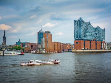 Hamburger Hafen mit Elbphilharmonie, Ausflugsbooten auf der Elbe und roten Backsteingebäuden unter bewölktem Himmel.