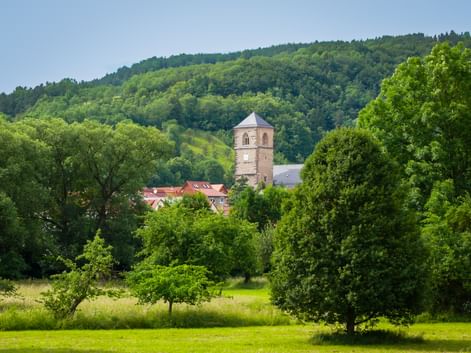Historic tower of Creuzburg Castle rising above red-roofed buildings, surrounded by lush green trees and forested hills in Thuringia.