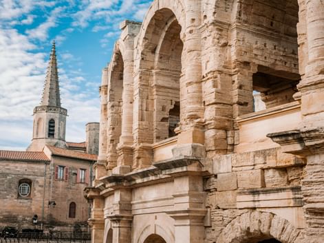 Ancient Roman amphitheater in Arles with weathered stone arches and columns. A church tower with red-tiled roofs visible in the background.