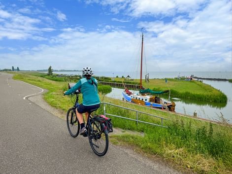 Cyclist in turquoise jacket riding on dike path alongside canal with moored sailboat. Green meadows and blue sky with clouds visible.