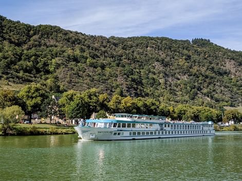 MS Switzerland Flusskreuzfahrtschiff fährt auf ruhigem grünen Wasser mit bewaldeten Hügeln im Hintergrund unter teilweise bewölktem Himmel.