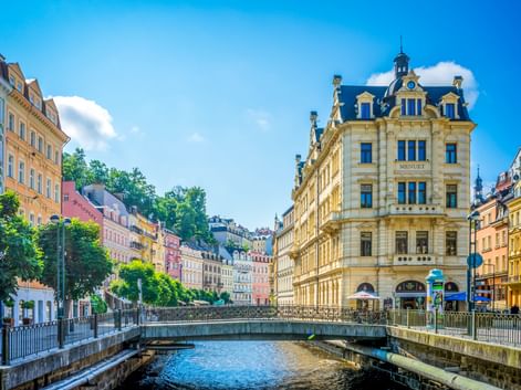 Historic Karlsbad old town with colorful buildings along the Tepla River. A yellow baroque building with blue roof stands prominently by the bridge.