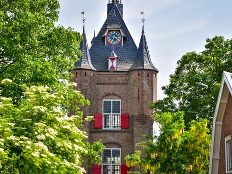 Historisches Stadttor Lekpoort in Vianen mit roten Backsteinmauern, Schieferdach, Uhr und roten Fensterläden, eingerahmt von grünen Bäumen.