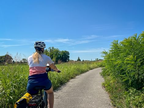 Radfahrerin mit blondem Pferdeschwanz und Helm auf asphaltiertem Weg durch grüne Wiesen mit hohen Gräsern und Farnen unter blauem Himmel.