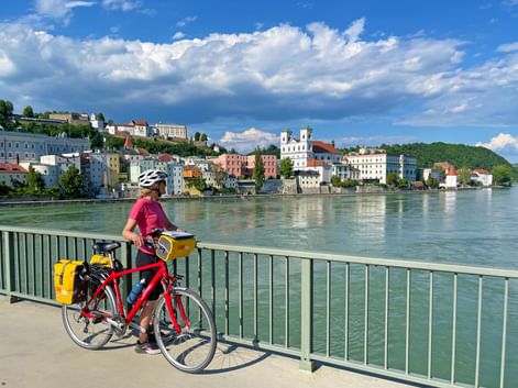 Radfahrerin mit rotem Fahrrad und gelben Packtaschen auf Brücke mit Blick auf Passauer Stadtbild entlang der Donau unter blauem Himmel.