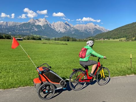Cyclist with child trailer on path through green meadow, Steinernes Meer mountain range in background under blue sky with white clouds.