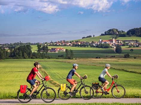 Three cyclists with panniers riding through green countryside near Michaelbeuern. Village with church visible on hillside in background.
