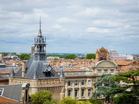 Aerial view of Toulouse with the Donjon du Capitole tower prominently featured. Red-tiled rooftops and historic buildings under a cloudy sky.