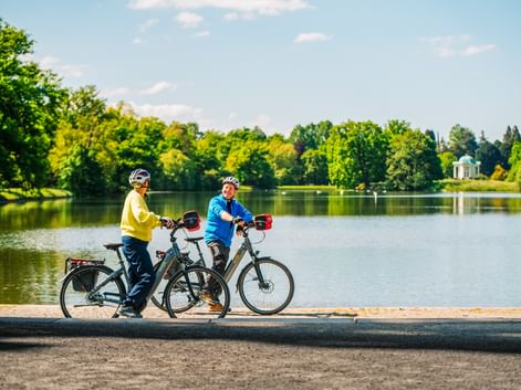 Two cyclists with e-bikes pause by a lake in Karlsaue Park, Kassel. A white pavilion is visible across the water surrounded by green trees.