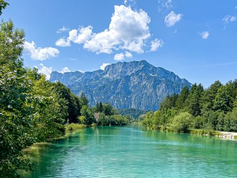 Turquoise Königsseeache river near Anif with rocky mountains in the background, framed by green trees under a blue sky with white clouds.