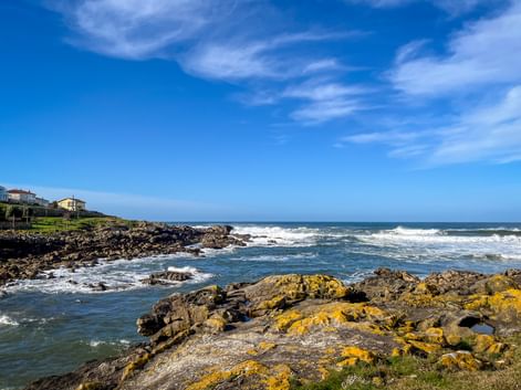 Rocky coastal landscape in A Guarda with yellow lichen-covered rocks, blue ocean waves, and houses on hillside under clear blue sky.