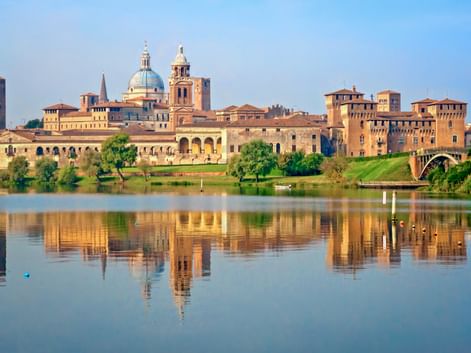 Historische Stadtansicht von Mantua mit Domkuppel und Glockenturm, die sich im ruhigen Seewasser spiegeln. Grüne Rasenflächen und Bäume säumen das Ufer.