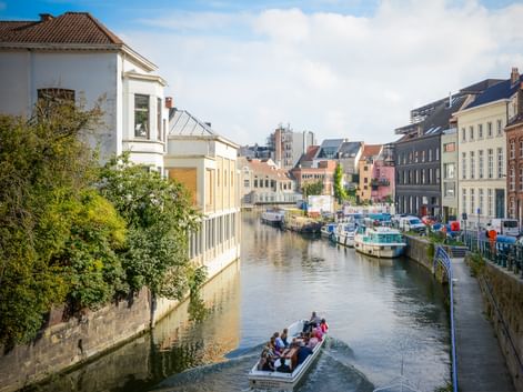 Canal in Ghent with colorful buildings, moored boats, and tourists on a boat tour. Trees line the waterfront under a blue sky with clouds.