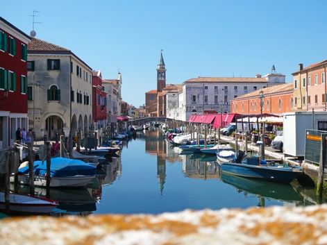 Kanal in Chioggia mit vertäuten Booten und bunten Gebäuden. Ein Kirchturm erhebt sich im Hintergrund unter blauem Himmel.