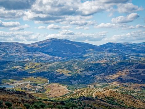 Weitläufige Berglandschaft bei Zaros, Kreta, mit sanften Hügeln voller Olivenhaine und landwirtschaftlichen Feldern unter blauem Wolkenhimmel.