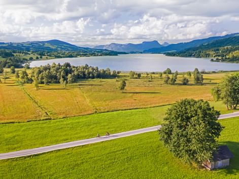 Two cyclists on a country road through green meadows with Irrsee lake and mountains under cloudy sky in Austrian Seenland region.