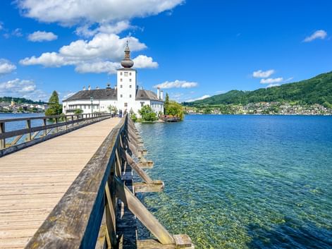 Holzsteg führt zu Schloss Orth am Traunsee im Salzkammergut. Klares blaues Wasser, weißes Schloss mit Turm, grüne Hügel.