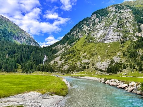 Turquoise Krimml Ache river flowing through green alpine meadow with forested mountains and rocky peaks under blue sky.