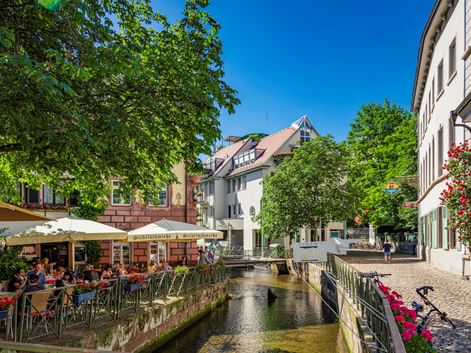 Historic Gerberau district in Freiburg with narrow canal flowing between traditional buildings. Outdoor café with umbrellas on left, flower-decorated buildings on right.