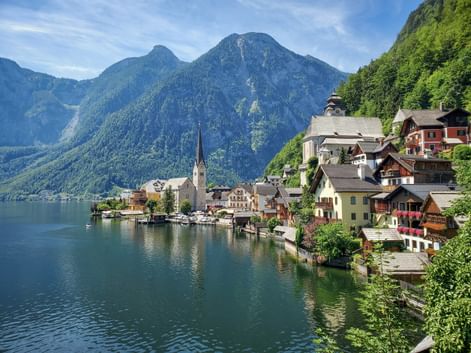 Hallstatt mit traditionellen Häusern und Kirchturm am Ufer eines türkisfarbenen Sees, umgeben von grünen bewaldeten Bergen.