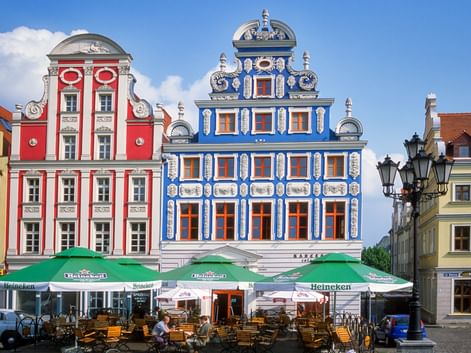 Red and blue ornate baroque buildings with white details at Sloneczny Plac in Szczecin. Green Heineken umbrellas shade outdoor café seating.