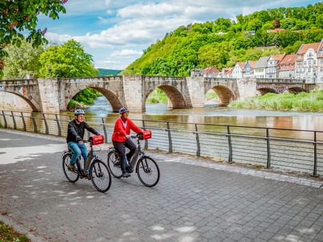 Zwei Radfahrer fahren entlang der Weser in Hann. Münden mit der historischen Werrabrücke und Fachwerkhäusern am Hang.