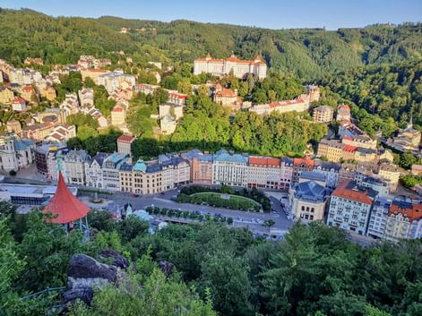 Aerial view of Karlsbad spa town with colorful historic buildings along a central square, surrounded by forested hills and green valleys.