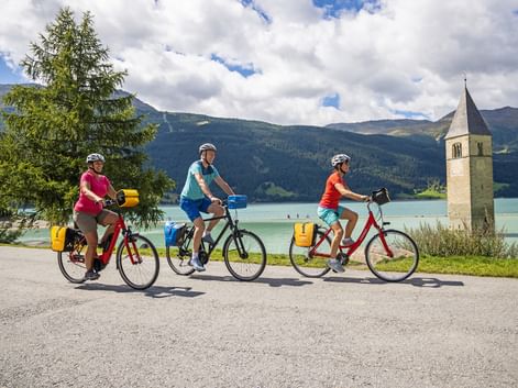 Three cyclists with yellow panniers riding along Lake Reschen. The famous church tower rises from the turquoise water, with mountains in the background.