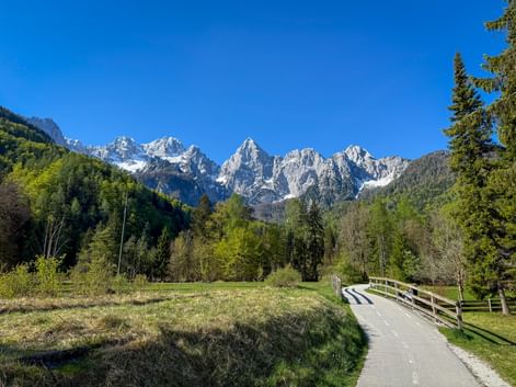 Paved cycling path with wooden bridge leading through green meadow toward snow-capped Julian Alps peaks near Kranjska Gora under clear blue sky.