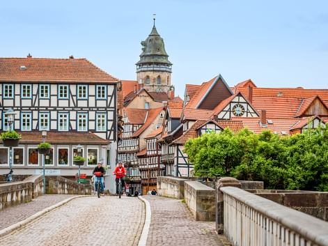 Zwei Radfahrer überqueren eine Steinbrücke zur Altstadt von Hann. Münden mit Fachwerkhäusern und Kirchturm unter klarem blauen Himmel.