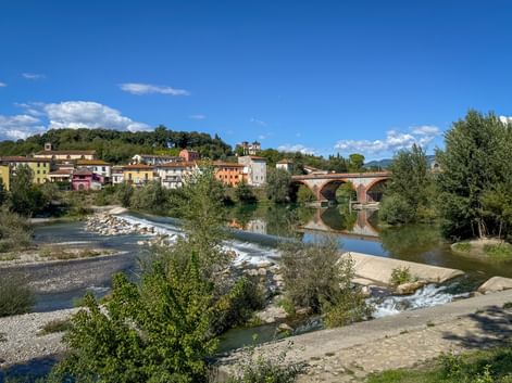 Stone bridge with arches spanning a river in Tuscany. Colorful buildings line the hillside behind the bridge, with trees and rocky riverbed in foreground.