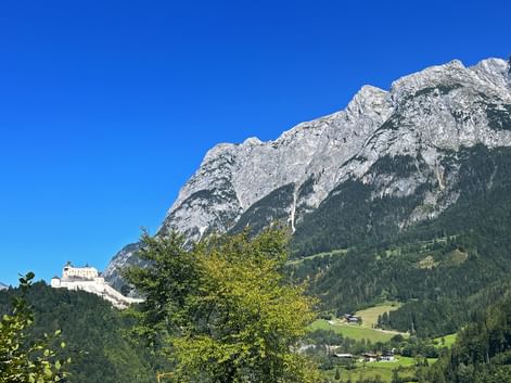 Burg Werfen auf einem bewaldeten Hügel mit den dramatischen Felsgipfeln des Tennengebirges dahinter unter klarem blauen Himmel.