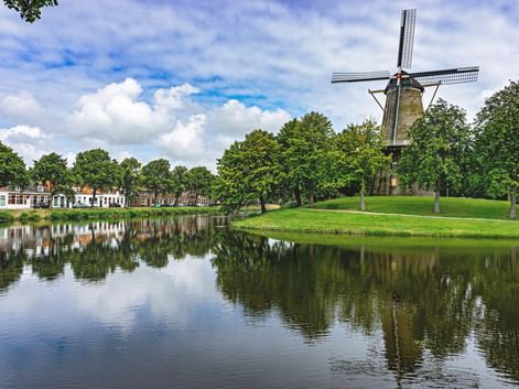 Traditional Dutch windmill Molen de Hoop on green lawn beside calm canal reflecting trees and houses in Middelburg under blue sky.