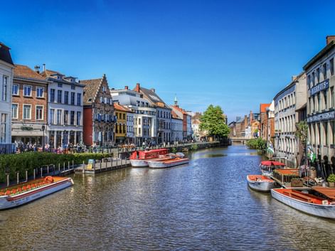 Kanal in Gent gesäumt von bunten historischen Gebäuden und Ausflugsbooten. Menschen sitzen am Ufer unter blauem Himmel.