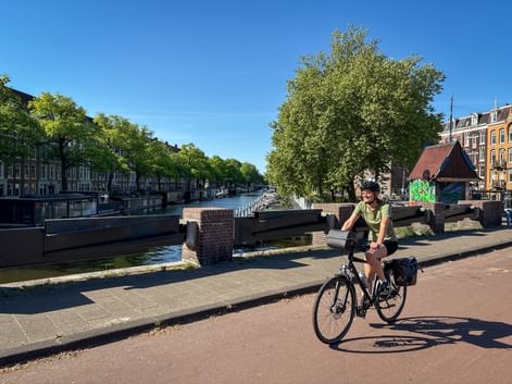 Radfahrerin fährt entlang eines Amsterdamer Kanals mit historischen Gebäuden und Bäumen am Wasser unter blauem Himmel.