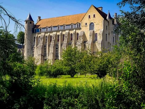 Historic Abbaye Saint-Séverin in Château-Landon with stone walls, arched windows, and round towers, surrounded by green meadow and trees.