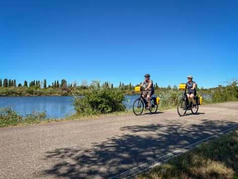 Zwei Radfahrer mit gelben Taschen fahren auf einem asphaltierten Weg neben einem Fluss in der Po-Ebene, mit Zypressen und blauem Himmel im Hintergrund.