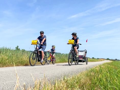 Family cycling on paved path with two adults on e-bikes with yellow panniers, child on bike, and bike trailer. Green fields under blue sky.