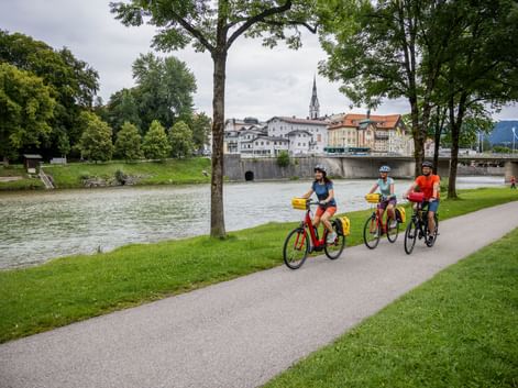 Drei Radfahrer fahren entlang der Isar in Bad Tölz. Historische Stadt mit Kirchturm und Brücke über dem Wasser, eingerahmt von grünen Bäumen.