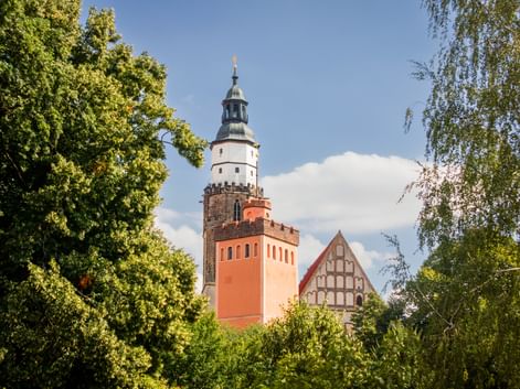 Roter Turm mit weißem Oberteil und dunkler Spitze neben Fachwerkkirche in Kamenz, eingerahmt von grünen Bäumen unter blauem Himmel.