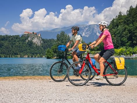 Two cyclists with panniers riding along Lake Bled shore. Bled Castle on cliff and mountains visible in background under blue sky with clouds.
