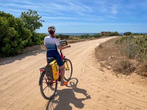 Cyclist with helmet and yellow panniers on sandy coastal path in Alto Minho, Portugal. Ocean visible in background under blue sky.