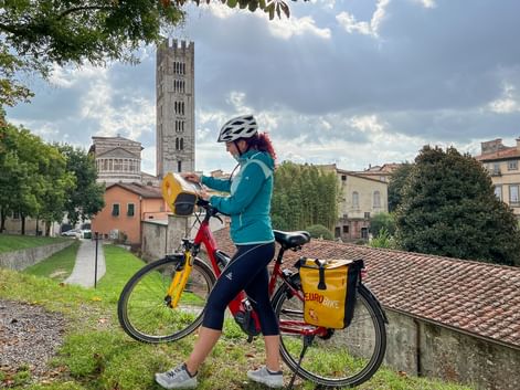 Female cyclist with yellow panniers on red bike in Lucca, Tuscany. Behind her rises a tall medieval bell tower against a cloudy sky.