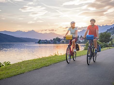 Two cyclists riding along lakeside path at Millstätter See during sunset, with mountains and village in background.