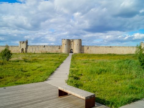 Wooden boardwalk leading through green marshland to the fortified walls of Aigues-Mortes with towers and gates under a cloudy sky.
